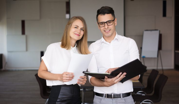 Portrait of happy male and female colleagues. Young Caucasian businessman wearing glasses standing with folder together with smiling female coworker in boardroom. Team concept